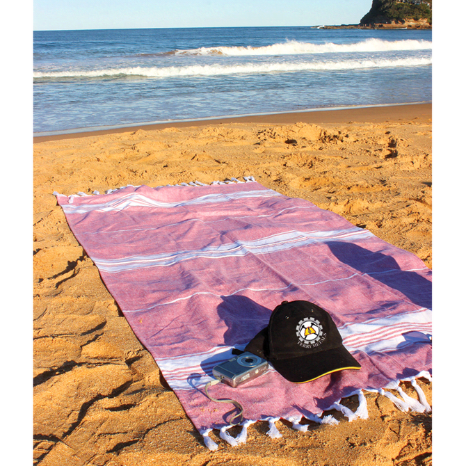 A pink and white striped Turkish towel laid on sandy beach, accompanied by a black cap and a compact camera.