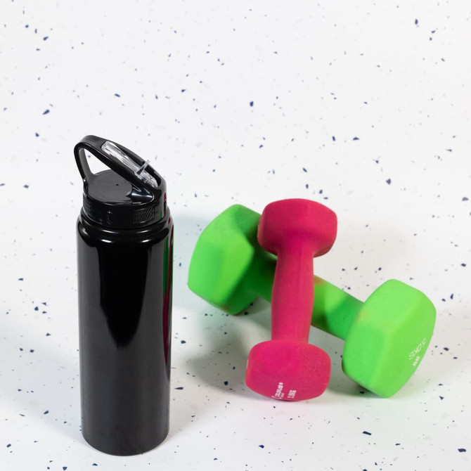 A drink bottle in sleek black with a flip straw lid, positioned beside pink and green dumbbells on a speckled background.