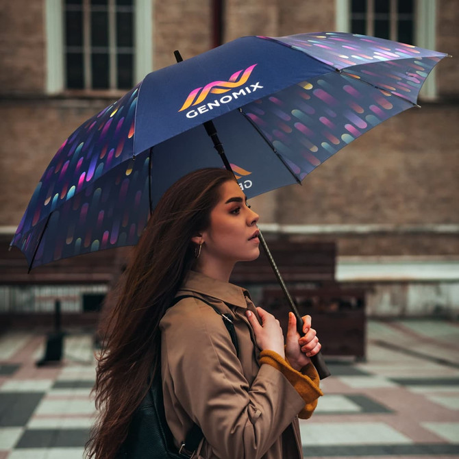 A full-colour umbrella in dark tones with a vibrant pattern, held by a woman with long hair, featuring a logo.