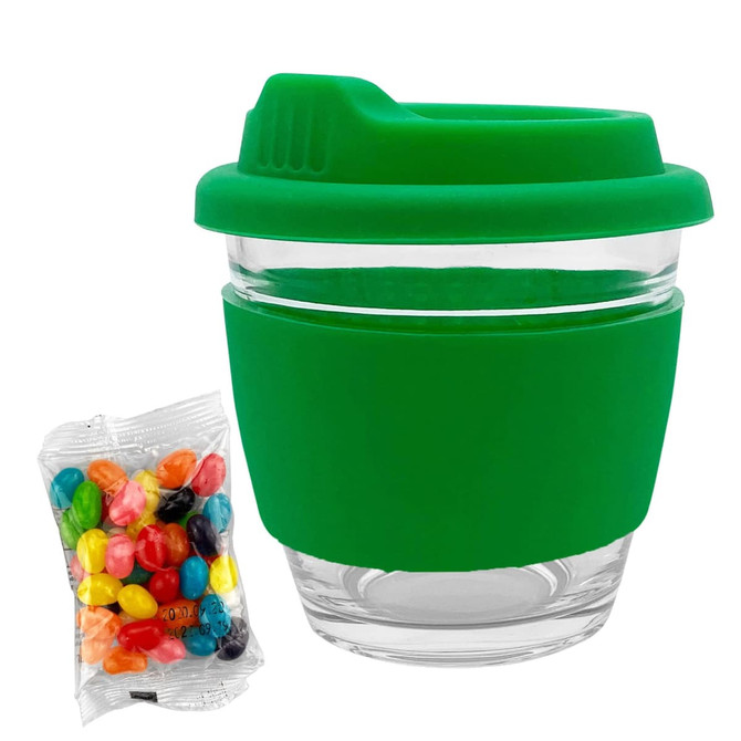 A reusable glass coffee cup with a green silicone sleeve and lid, accompanied by a bag of colourful jelly beans.
