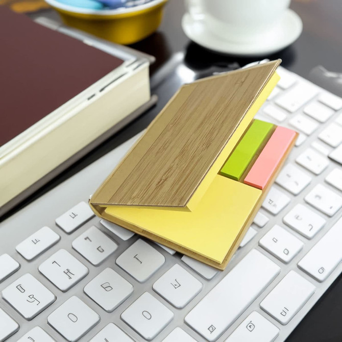 A bamboo sticky note holder with yellow, green, and pink sticky notes, placed on a white keyboard beside a book and a cup.