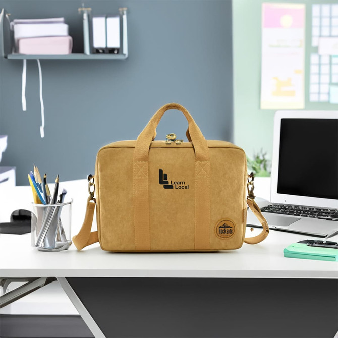 A yellow kraft paper satchel with handles and a shoulder strap, featuring a logo, sitting on a desk.