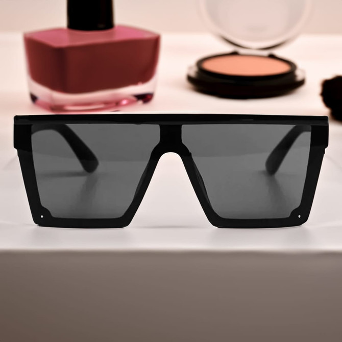Black oversized sunglasses with dark lenses, displayed in front of cosmetics on a flat surface.