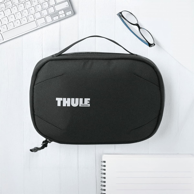 A black toiletry bag with a handle and a logo on the front, placed on a light wooden surface with office supplies.