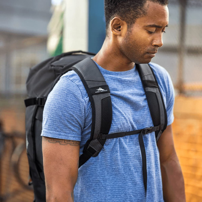 A man wearing a blue shirt carries a black duffel backpack, showcasing its straps and design details.