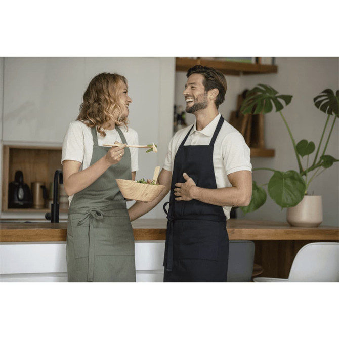 A green and a navy apron worn by two people laughing together in a kitchen setting. The scene features a wooden bowl of salad.