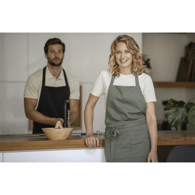 A green apron is worn by a smiling woman in a kitchen, while a man in a black apron stands behind her.