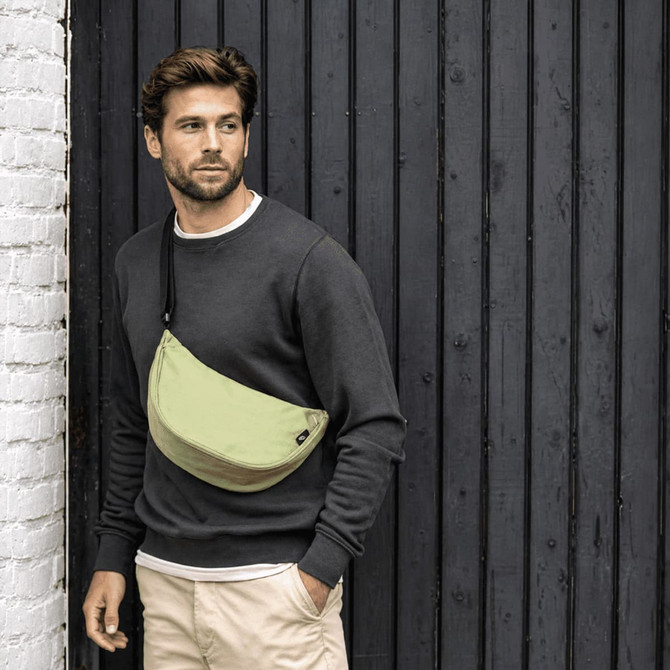 A waist bag in light green worn across a man's chest, with a casual grey sweater and beige pants, against a dark wooden background.