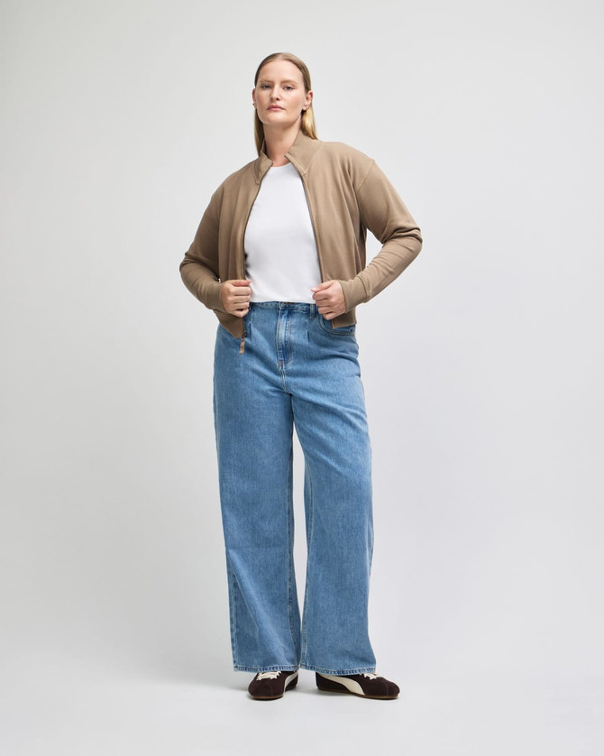 A woman wears a beige full-zip jacket over a white top, paired with wide-legged blue jeans. The jacket features a logo.