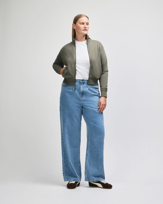 A woman wearing an olive green full-zip jacket over a white t-shirt, paired with light blue wide-leg jeans.