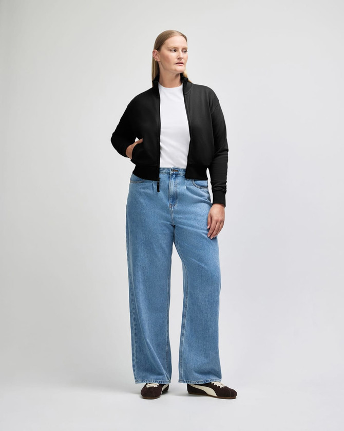 A woman wearing a black full-zip jacket and blue jeans, standing with one hand on her hip. The jacket has a logo.