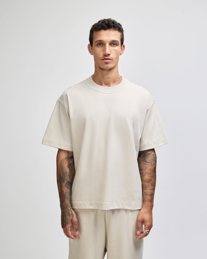 A unisex boxy t-shirt in a light beige colour, worn by a model with tattoos against a plain background.