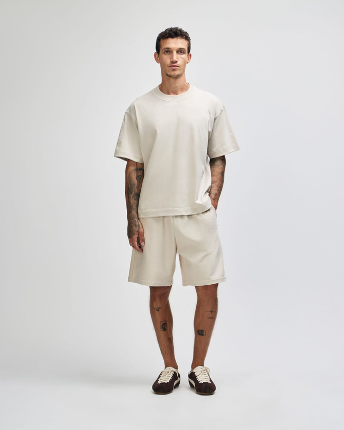 A model wears a light beige heavyweight cotton unisex boxy t-shirt and matching shorts, featuring a logo.