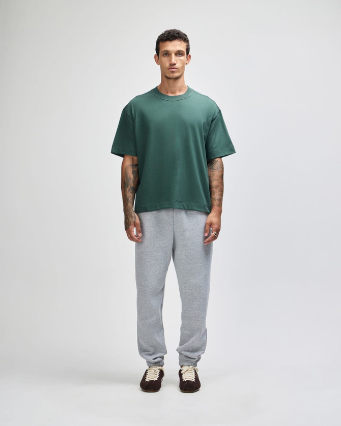 A unisex boxy t-shirt in green, paired with grey sweatpants, worn by a model standing against a plain background.