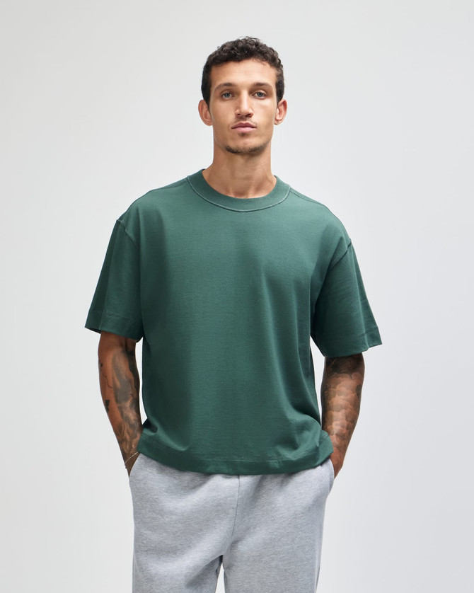 A green heavyweight cotton unisex boxy t-shirt, worn by a model with short hair, paired with grey sweatpants.
