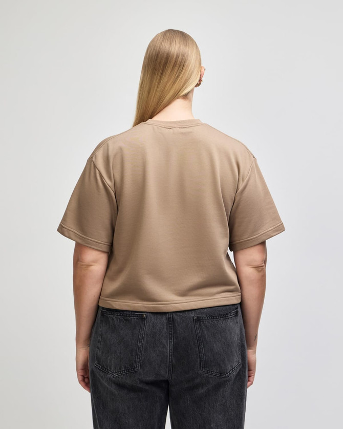 A back view of a beige mid-length boxy top with short sleeves, paired with dark grey pants. The top features a logo.