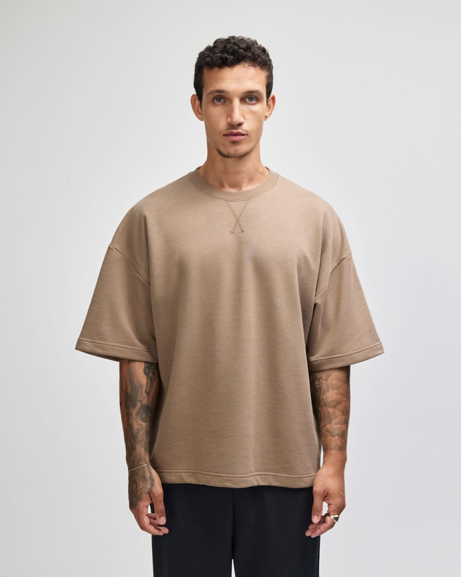 A unisex oversized boxy top in a tan colour, featuring a casual fit and short sleeves. It has a visible logo.