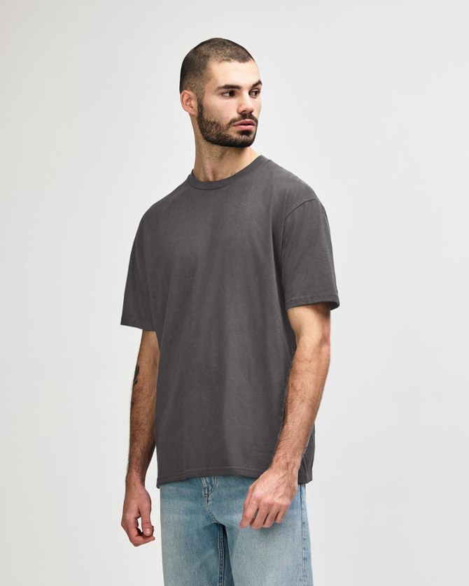 A man stands wearing a midweight T-shirt in olive green. The shirt features a logo on it.