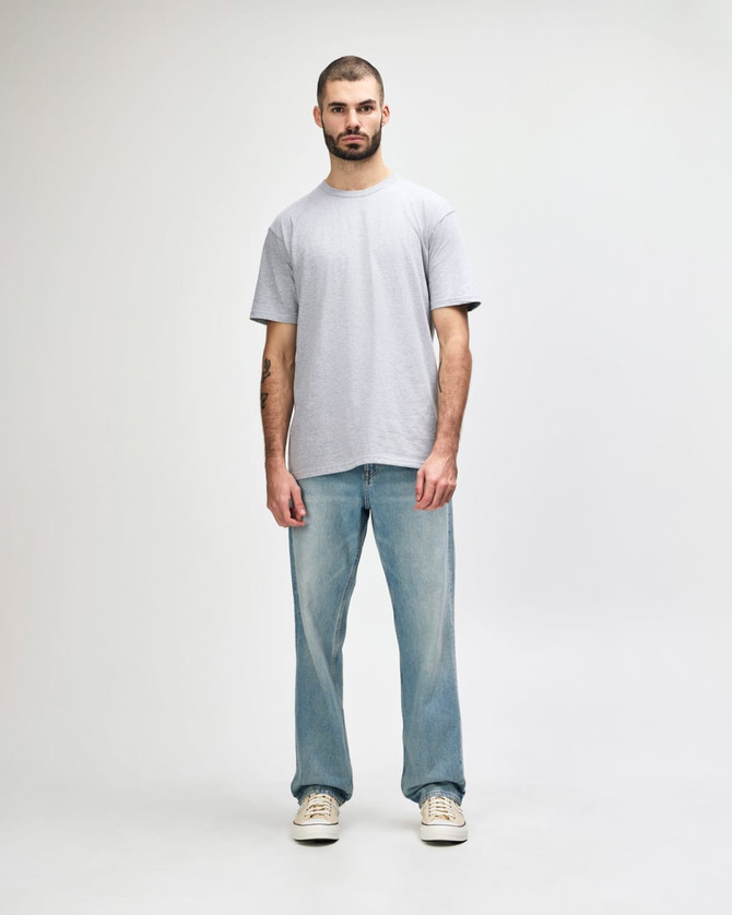 A man stands wearing a light grey Gildan Softstyle adult midweight T-shirt and blue jeans. The shirt has a logo.