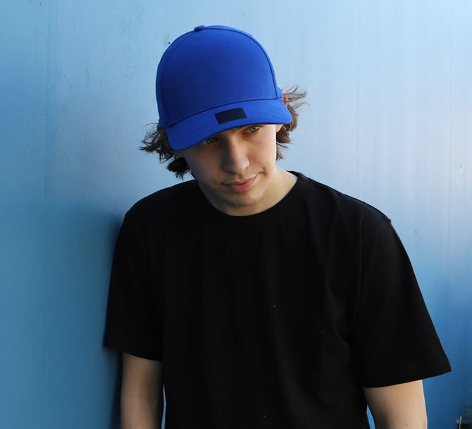 A blue cotton and spandex cap worn by a person in a black t-shirt, leaning against a blue wall.