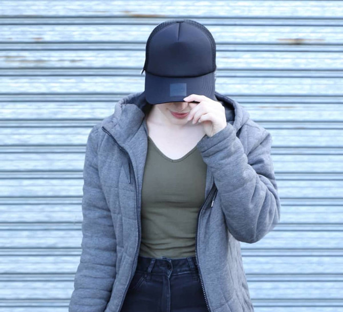 A black trucker cap with mesh detailing, worn by a person in a grey jacket and olive green top, standing against a striped background.