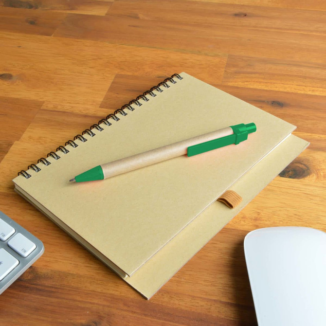 A brown spiral notebook placed on a wooden desk with a green pen resting on top and a computer mouse nearby.