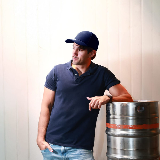 A young man wearing a navy blue polo shirt and cap leans against a silver beer keg.