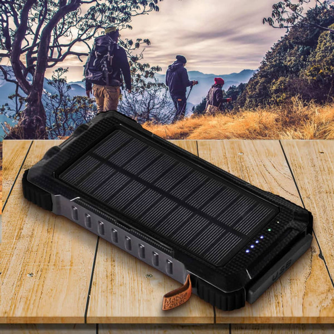 A black solar power bank with a textured surface and LED indicators, placed on a wooden table in a scenic outdoor setting.