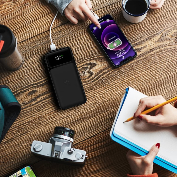 A black wireless power bank with a digital display charging a smartphone, surrounded by a camera, notebook, and coffee cup.