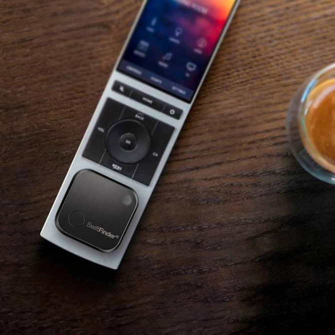 A black SwiftFinder Smart Tag Tracker on a remote control, placed on a wooden surface beside a coffee cup.