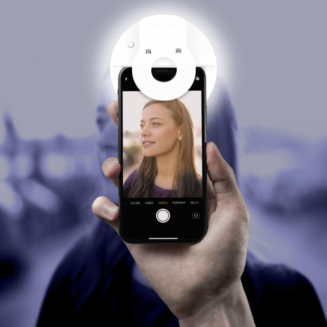 A hand holds a smartphone with a circular white Smiley LED Selfie Light attached, illuminating a person's face in the photo.