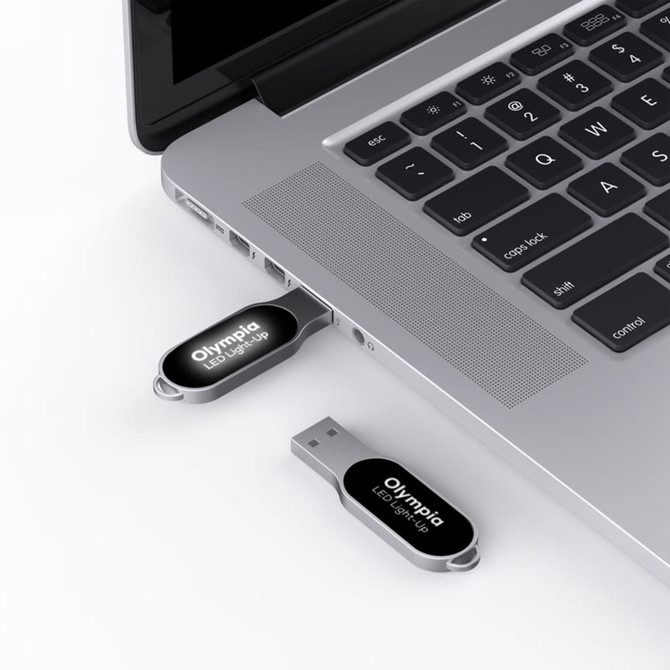 A silver Olympia LED flash drive is plugged into a laptop's USB port, with another flash drive beside it.
