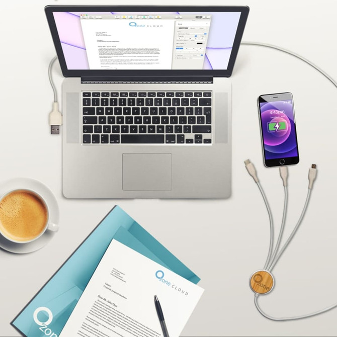 A laptop, a smartphone charging with a gold Murano Charge Cable, a coffee cup, and stationery on a desk.