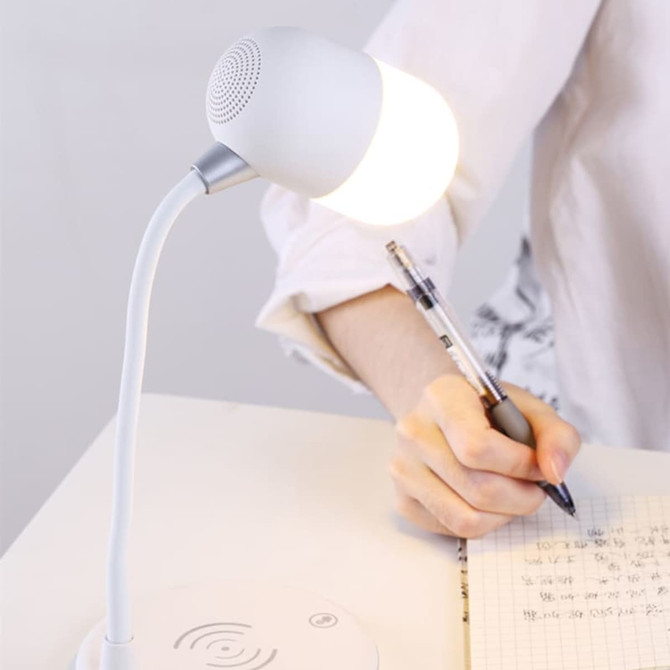 A white wireless charging sound lamp with a curved neck and glowing bulb sits on a desk beside a person writing.