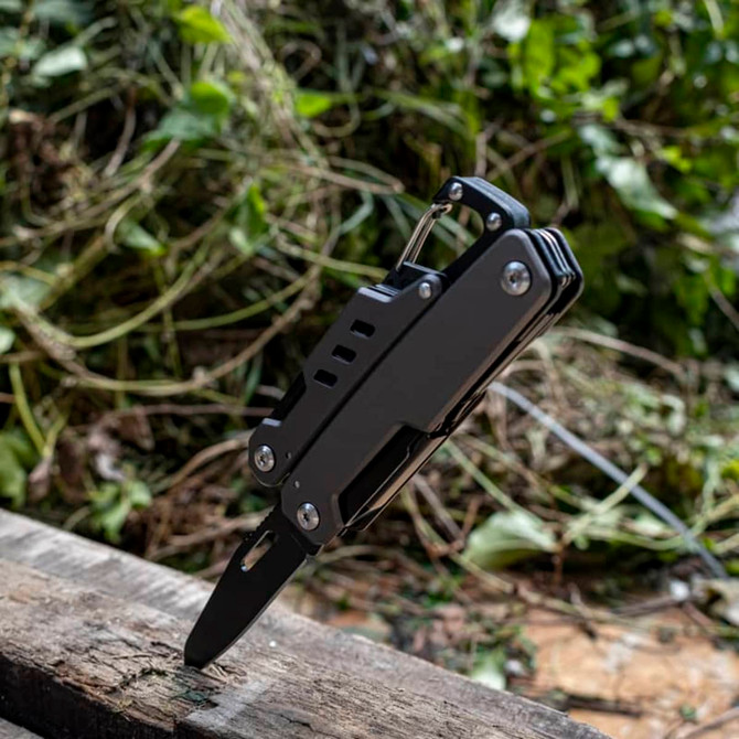 A black multi-tool featuring various tools, including a knife, stably positioned on a wooden surface amidst foliage.