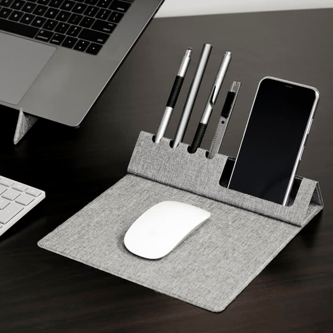 A grey foldable mouse pad with a phone holder and slots for pens, placed beside a laptop and keyboard.