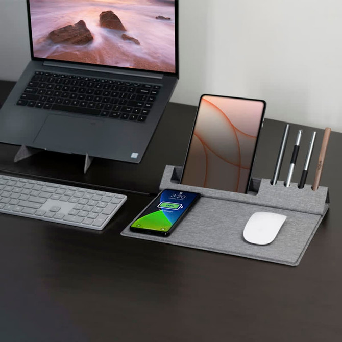 A grey eco mouse pad with a phone stand, holding pens and a smartphone, placed on a desk next to a laptop and keyboard.