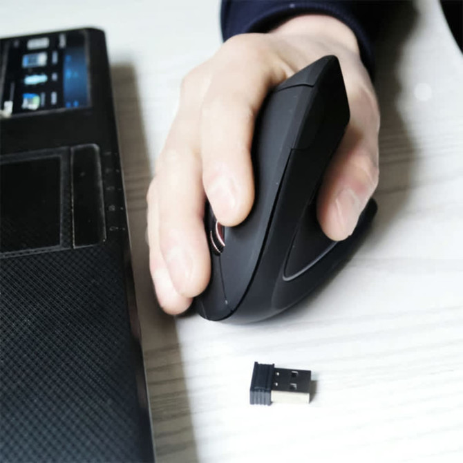 An ergonomic wireless mouse in black held in a hand, positioned beside a laptop with a USB receiver nearby.