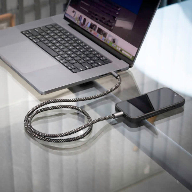 A grey and black charging cable connected to a smartphone and a laptop on a glass table.