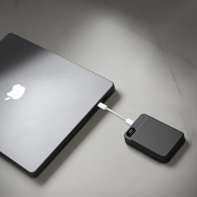 A black magnetic wireless power bank connected to a laptop on a light surface. The power bank has a logo.