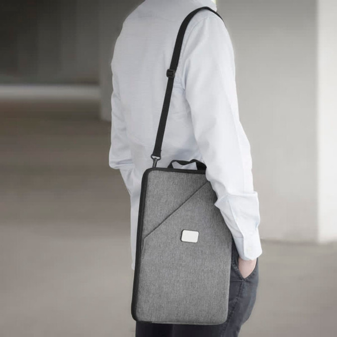 A grey laptop sling bag with a shoulder strap is held by a person wearing a light blue shirt. It features a branded logo.