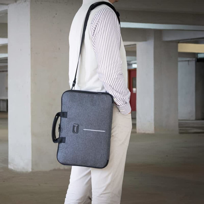 A grey laptop case with a shoulder strap and handle, shown being carried by a person in an indoor setting.