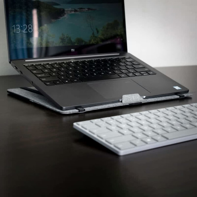 A grey laptop stand supports a laptop on a dark wooden desk, with a white keyboard nearby.