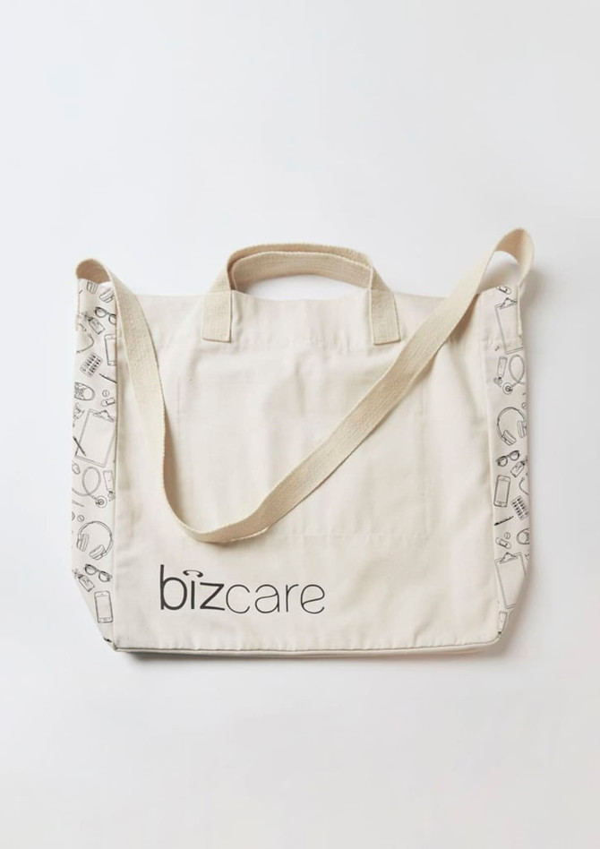 A cream tote bag with a strap, featuring a logo and patterned detailing along the edges.