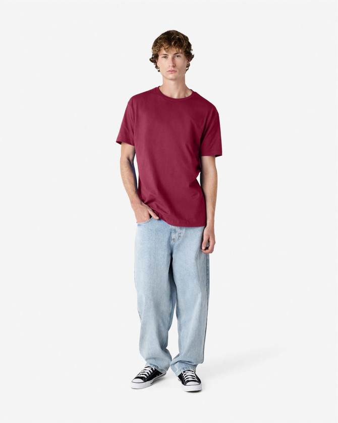 A unisex heavyweight cotton t-shirt in maroon, worn with light blue baggy jeans and black sneakers, featuring a logo.