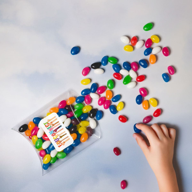 Assorted mini jelly beans in various colours spilling from a clear pillow pack, with a hand reaching for them.