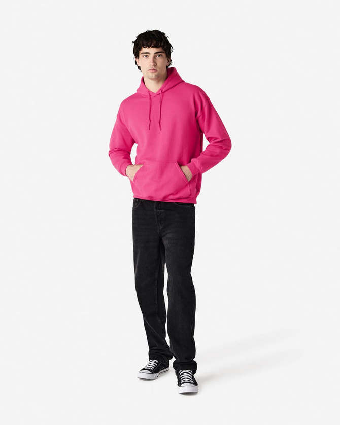 A man stands wearing a pink hooded sweatshirt with a front pocket, paired with black jeans and sneakers.