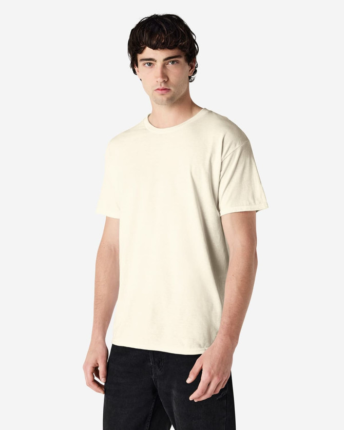 A man models a beige Gildan Heavy Cotton Adult T-Shirt, with a minimalistic design and short sleeves.