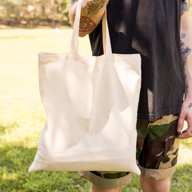 A short handle tote bag in natural beige is held by a person in a casual outfit, featuring a logo.