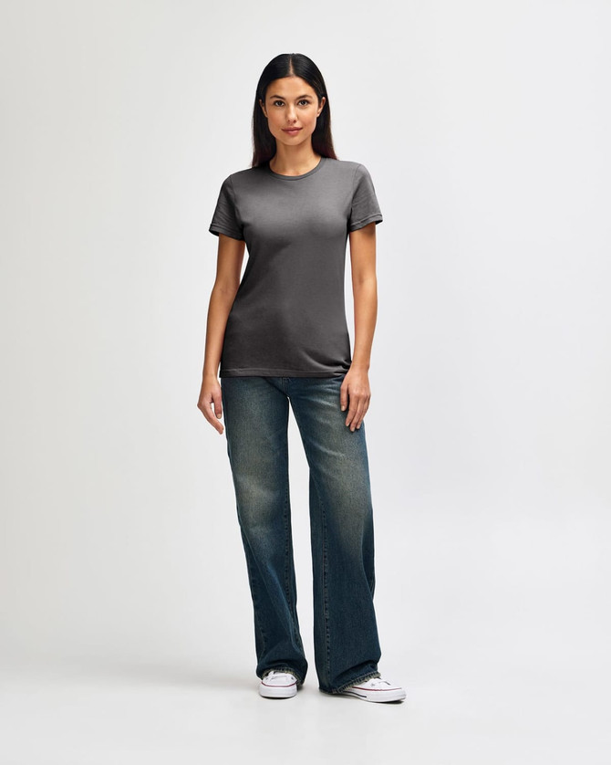 A woman stands wearing a soft-style ladies' t-shirt in grey, paired with blue jeans and white sneakers.